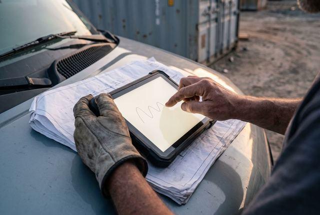 Zwei Hände unterschreiben mit dem Finger auf einem Tablet, das auf einer Bauzeichnung liegt.